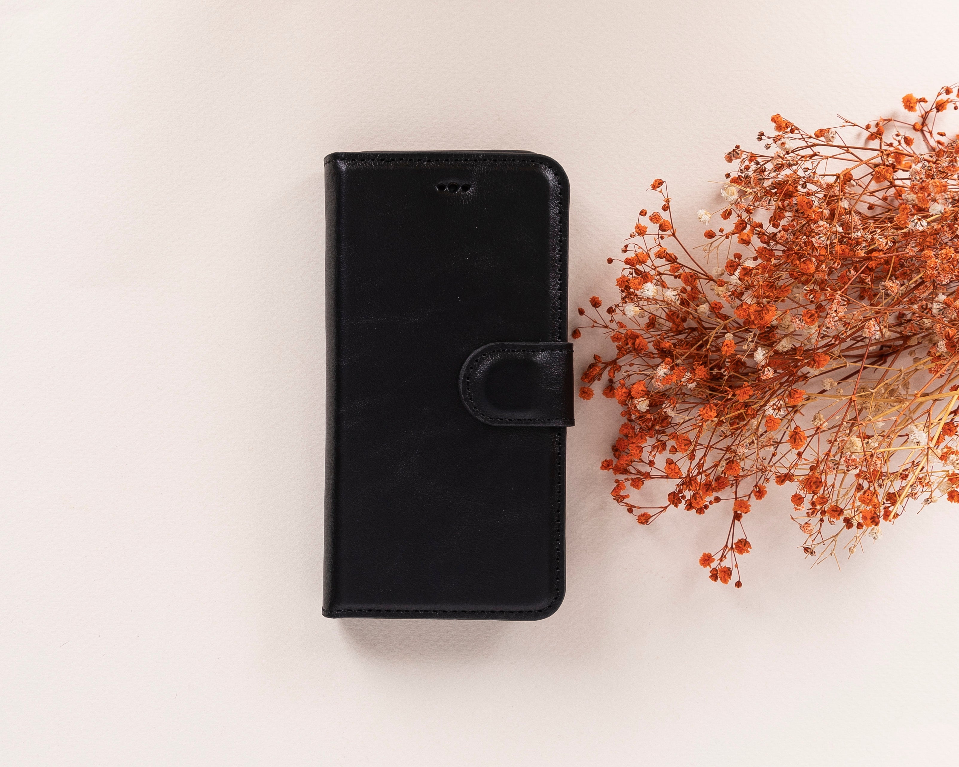 A black leather iPhone case with a detachable wallet feature, displayed alongside some dried orange flowers. The case has card slots visible on the front and a magnetic closure.