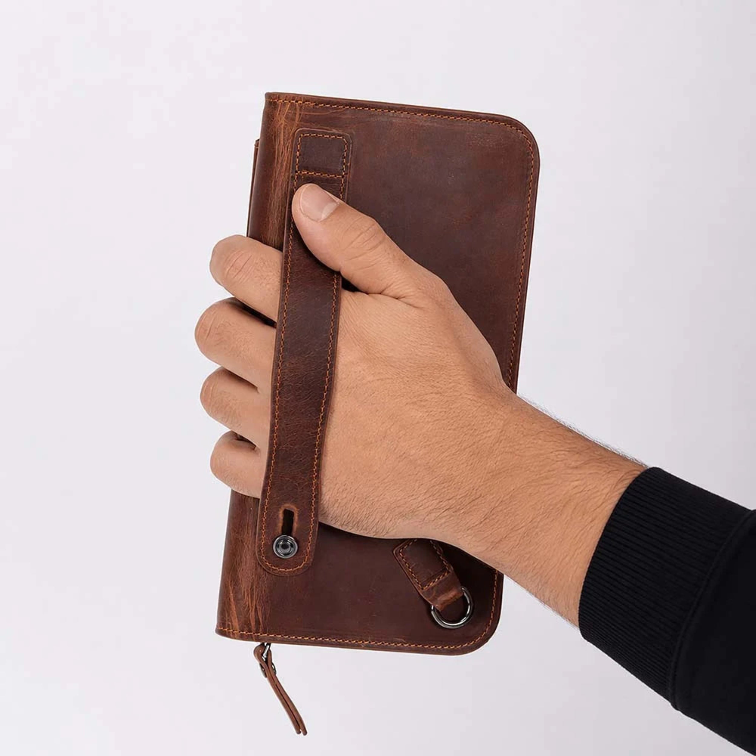 A hand holding a brown leather zip-around wallet or notebook cover with a wrist strap and metal snap button, shown against a plain light background.