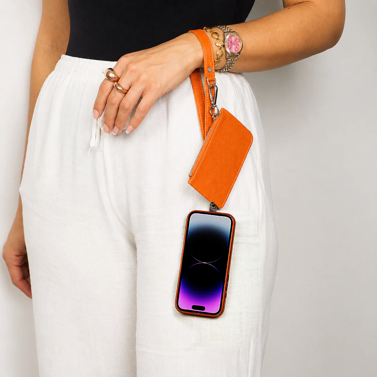 An orange leather phone strap and matching card holder pouch with a gold-tone clasp, worn around the waist of a model wearing white drawstring pants.