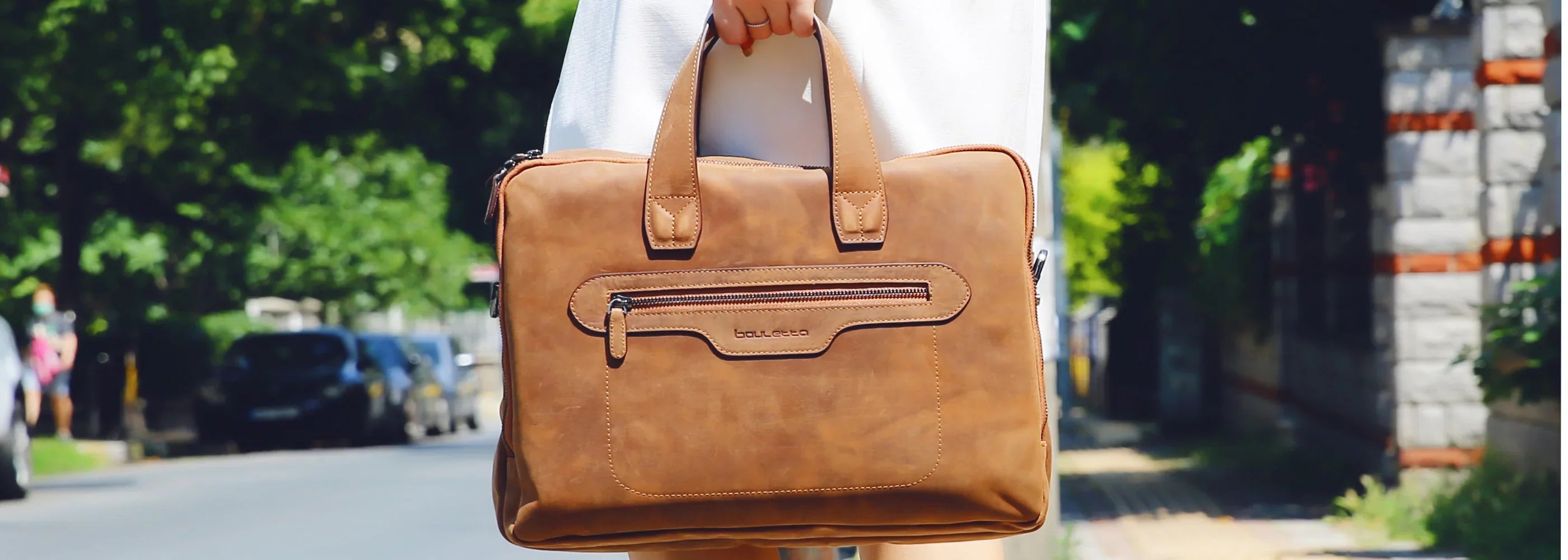 Person holding a brown leather briefcase outdoors on a sunny day