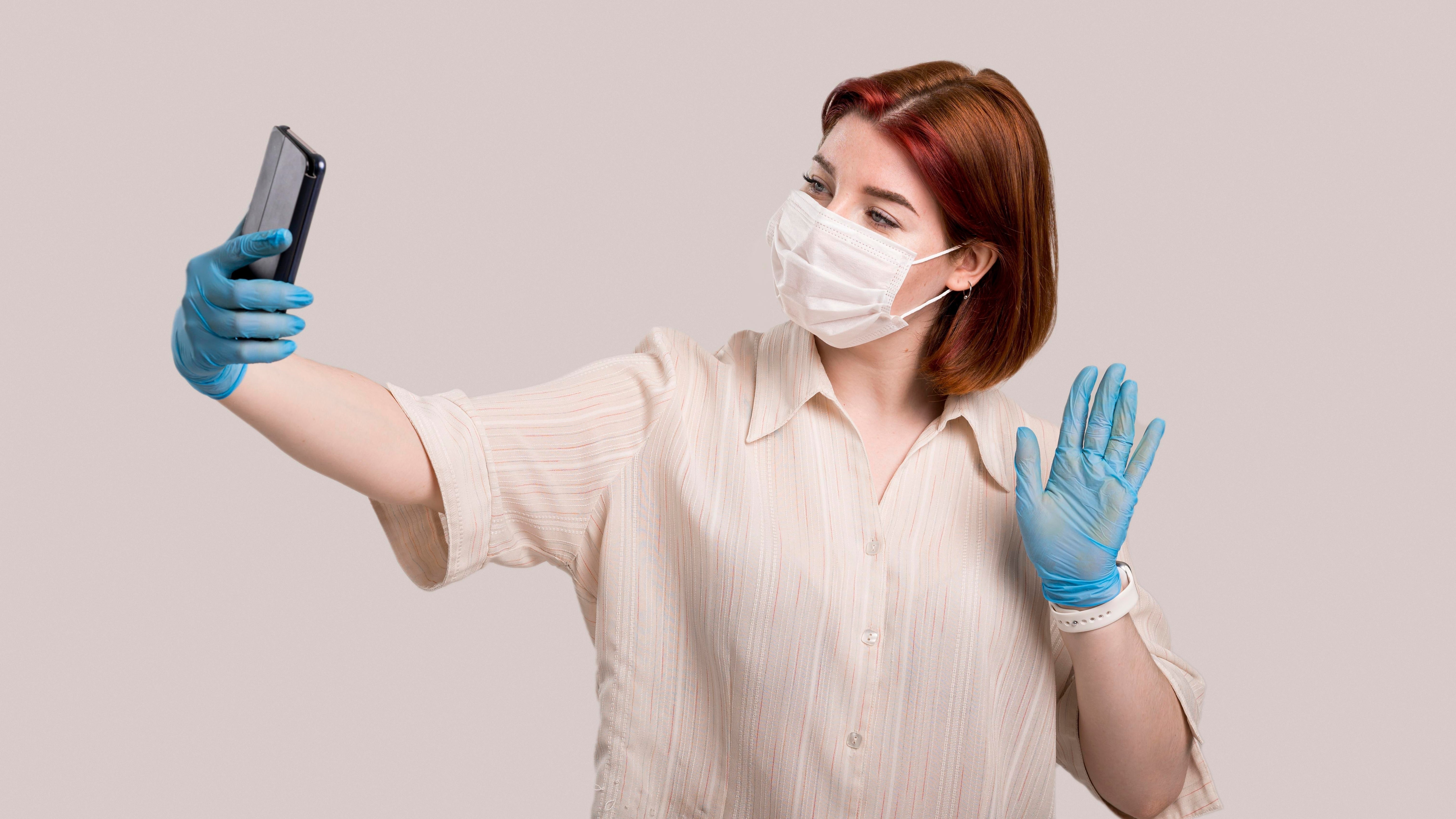 Woman wearing a face mask and blue gloves holding a smartphone and waving