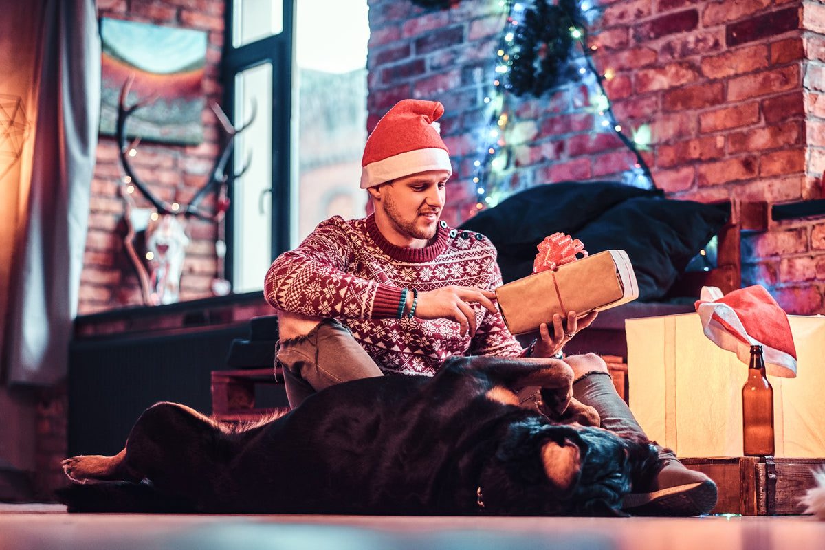Man in a Christmas jumper opening a leather gift beside his dog in a decorated living room