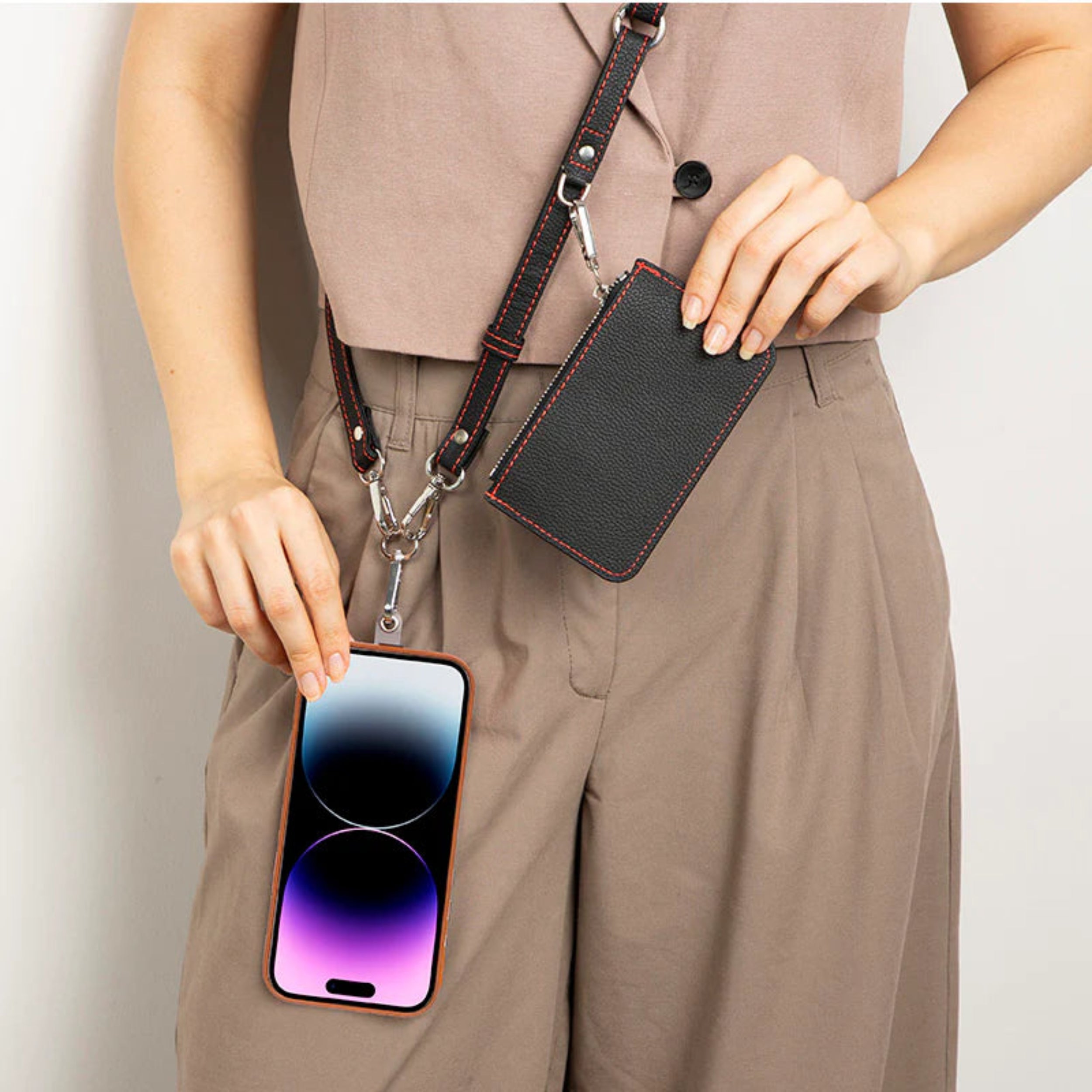 A person wearing a taupe top and matching pants demonstrates a black leather crossbody phone pouch with a brown leather strap and silver hardware. The pouch is shown holding a smartphone and a small card, with the phone and card hanging from the strap.