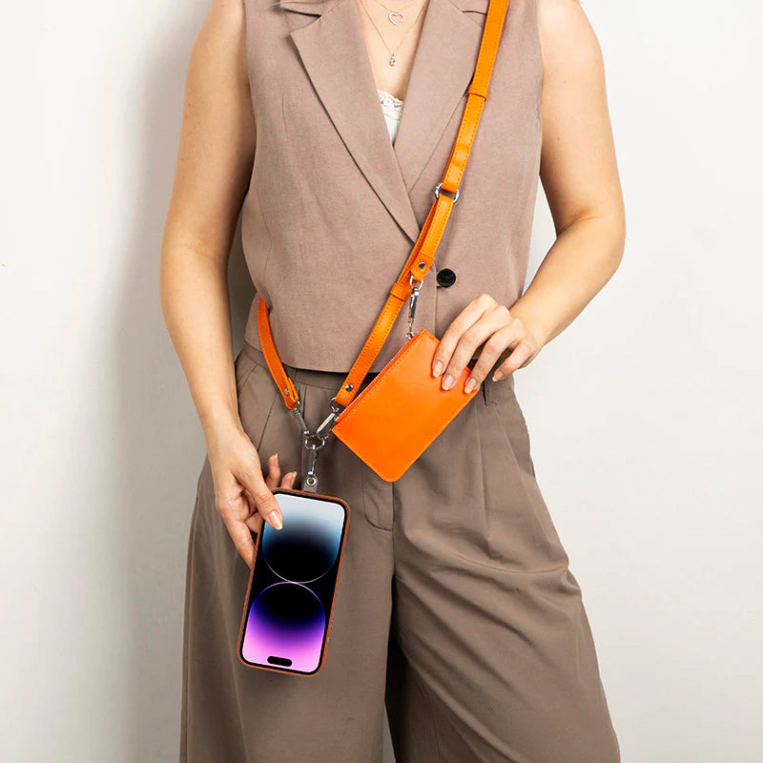 A person wearing a taupe blazer vest and matching trousers models an orange crossbody phone bag with a detachable strap and a smartphone hanging from the bag.