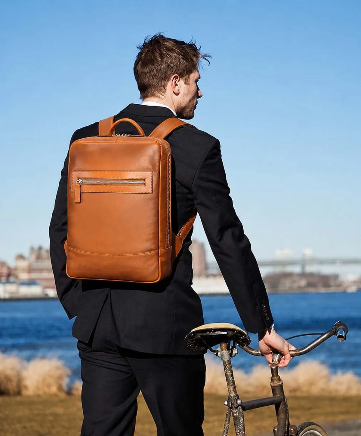 A man in a black suit seen from behind wearing a tan leather backpack, standing next to a vintage bicycle outdoors near the water under a clear blue sky.