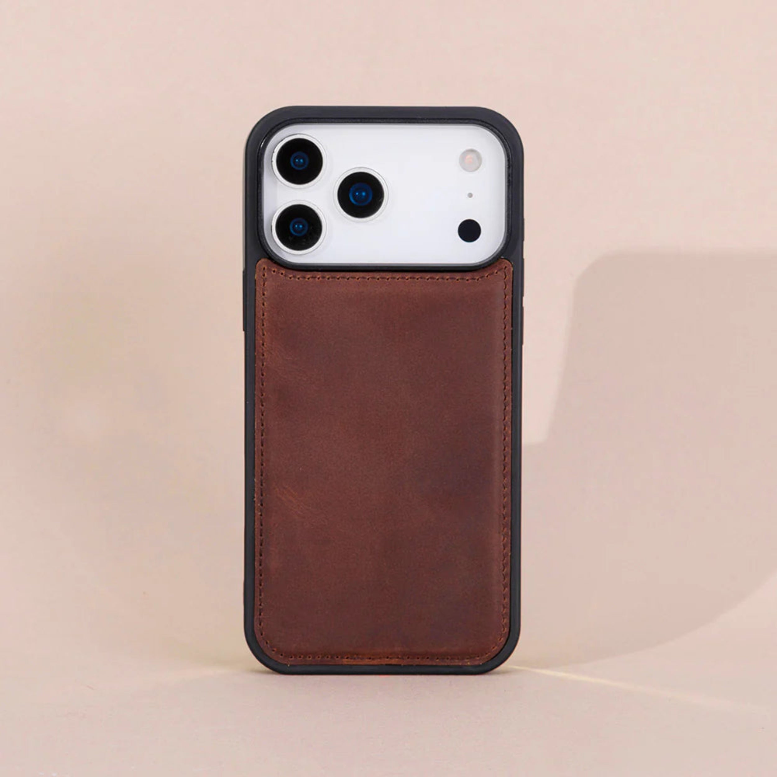 A brown leather phone case with a black bumper frame, shown on a white smartphone against a warm beige background.