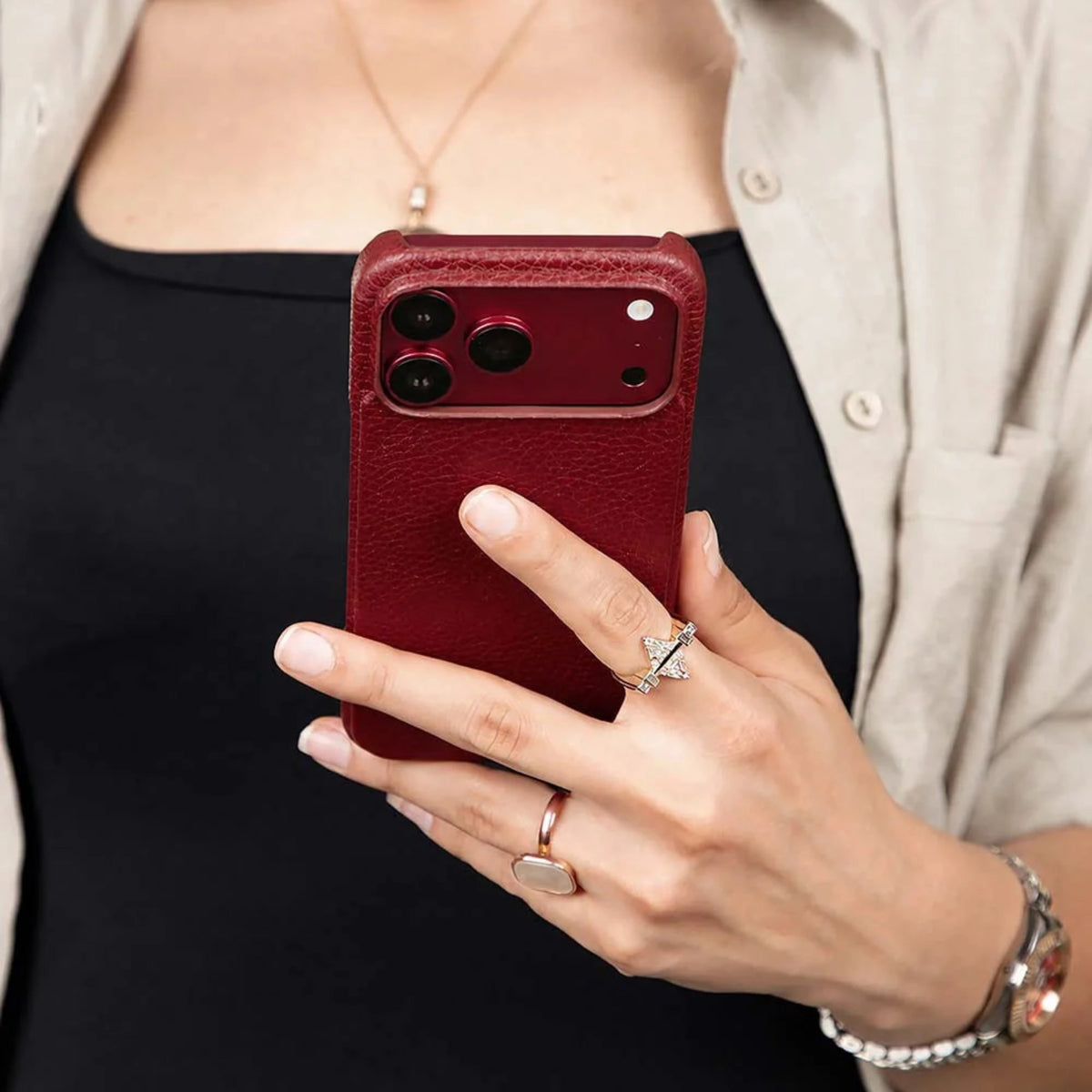 A close-up of a person holding a smartphone with a textured deep red leather case. The person is wearing a black top, a light beige button-up shirt, and several rings, including a silver bow ring on the index finger.