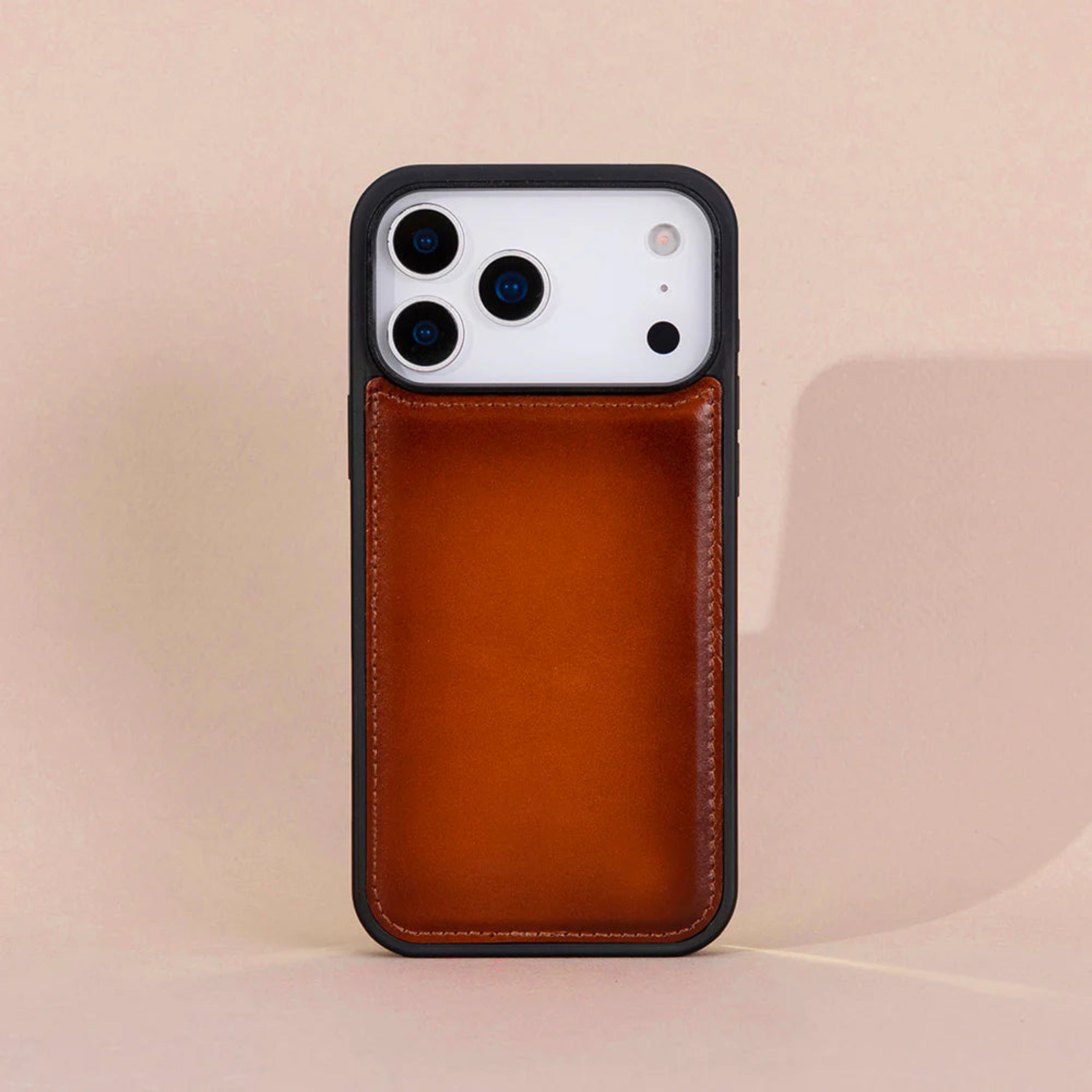 A iphone 17 pro case with a warm brown leather back panel and a black bumper edge, shown standing upright against a soft beige background.