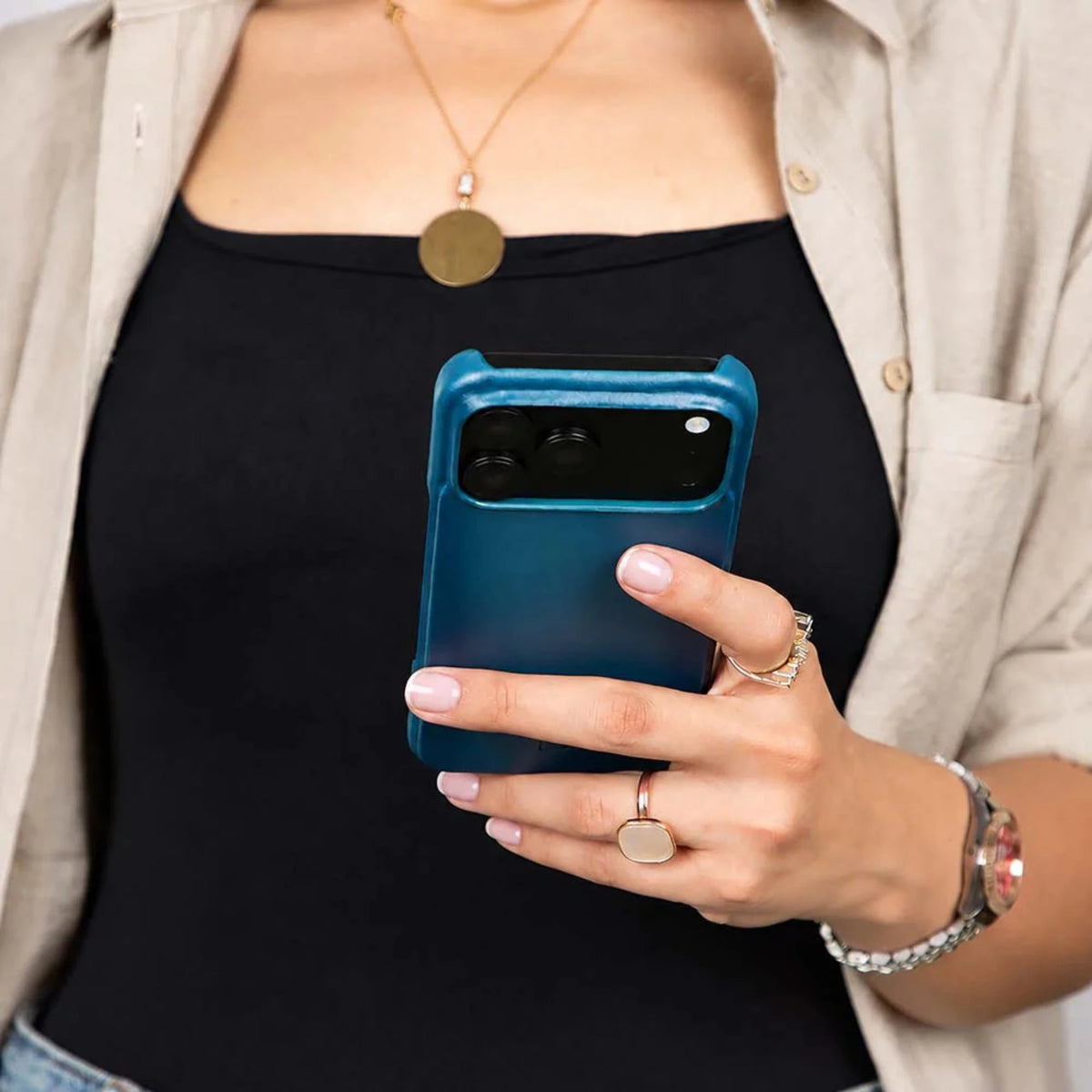 A close-up of a person holding a blue smartphone case with a black camera cutout, wearing a black top, beige button-up shirt, and gold jewelry.
